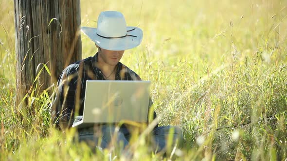 Cowboy sits in grass using laptop computer