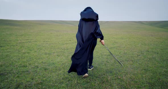 Witch Wearing Black Cape Is Walking in the Field, Holding a Sword, Halloween, 