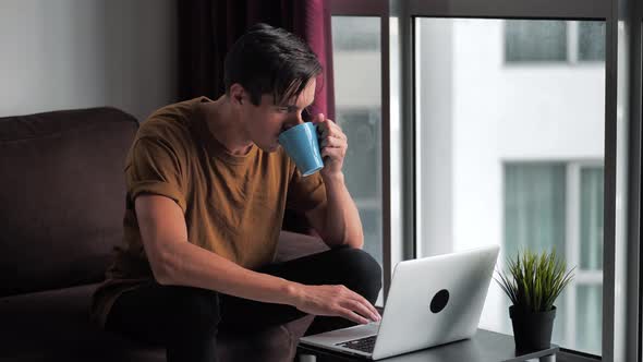 Handsome Young Man Drinking Coffee While Using A Laptop Computer Sitting at Home on a Sofa in Front