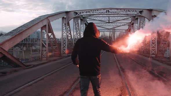 Young Man in Hoodie and Balaclava with Red Burning Signal Flare on the Road Under an Old Steel Frame