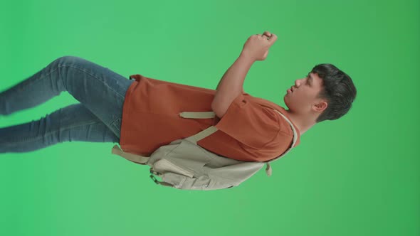 Side View Of Asian Boy Student Playing Game On Mobile Phone While Walking On Green Screen