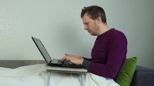 Young Unshaven Man Works at the Computer at Home Sitting in Bed. The Concept of Work at Home