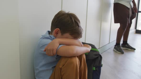 Video of sad caucasian boy sitting at school corridor, covering head with hands