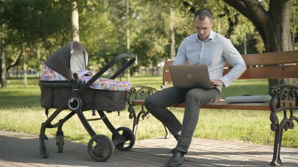 Wide Shot of Confident Busy Caucasian Man Surfing Internet on Laptop and Rocking Baby Carriage