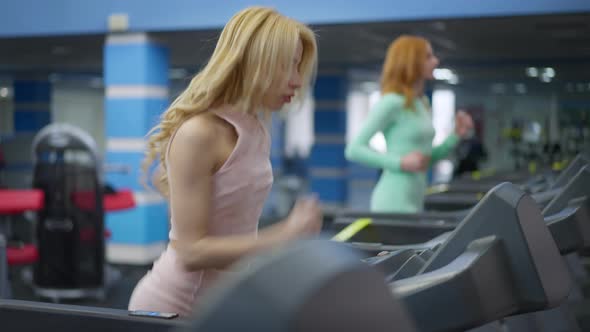 Medium Shot Portrait of Beautiful Young Caucasian Slim Woman Jogging on Treadmill As Blurred Redhead