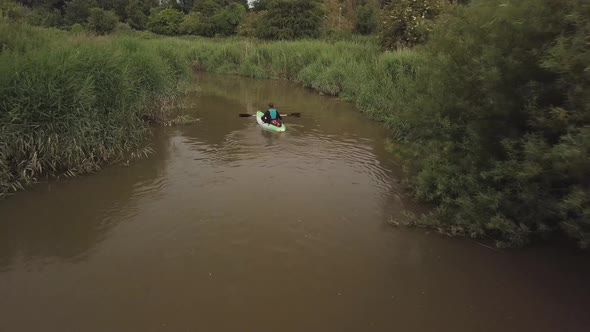 Man kayaking on meandering river with reeds bush’s and trees on either bank. Drone follows behind th