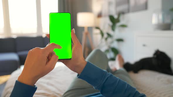 Woman at Home Lying on a Bed with Black Cat and Using Smartphone with Green Mockup Screen in