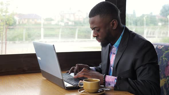 Black Businessman is Working Typing a Message on Laptop Sitting in Summer Cafe