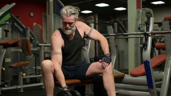 A gray-haired man with a beard and tattoos pumps biceps in the gym