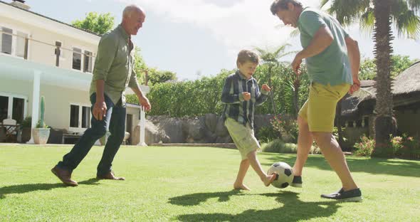 Happy caucasian grandfather with adult son and grandson playing football in garden