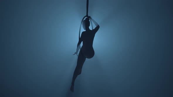 Silhouette of Flexible Aerial Gymnast Rotates on an Aerial Ring and Performs Acrobatic Tricks in the