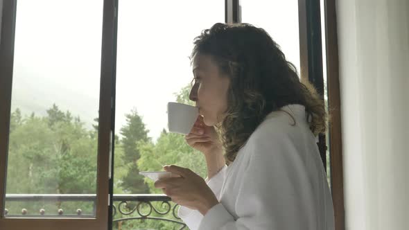 Young Beautiful Woman in a White Bathrobe Drinking Morning Coffee From a White Cup in the Room