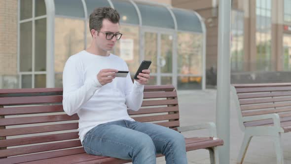 Young Man Reacting to Online Payment Failure on Smartphone While Sitting Outdoor on Bench