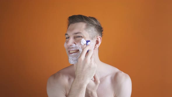 Man Shaves His Beard Using Foam and a Razor While Posing for the Camera Against an Isolated Orange