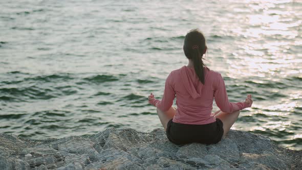 Young beautiful sport girl training meditation sitting on stones seaside while sunset.