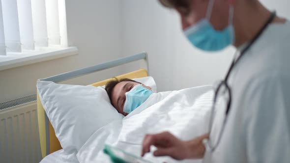 Patient Lying in Bed Talking to Doctor Entering Data in Digital Tablet Sitting in Hospital Ward