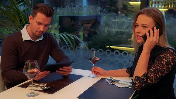 A Man and a Woman Sit at a Table in a Restaurant, He Works on a Tablet, She Talks on a Smartphone