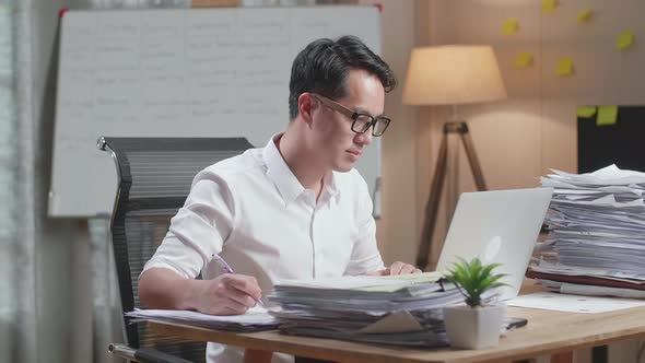 Asian Man Looking At Laptop And Taking Note While Working With Documents At The Office