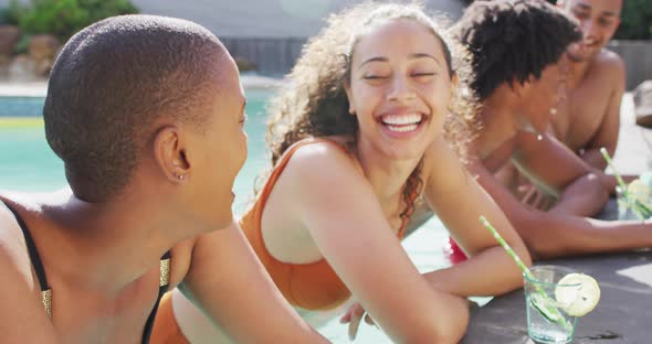 Two diverse female friends with drinks laughing by swimming pool