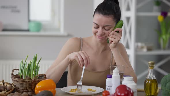 Portrait of Sad Slim Beautiful Young Woman Sitting at Table with Cucumber Mixing Supplement Pills