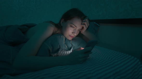 Young Caucasian Woman Lying in a Bed in the Bedroom Browsing the Internet Using a Smartphone Sleepy