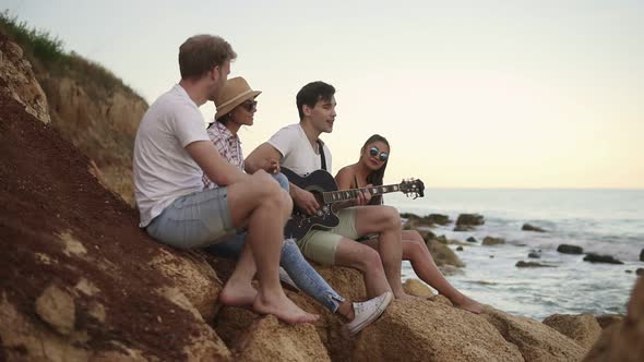 Group of Young Hipster Friends Sitting on the Rocks By the Seashore and Playing Guitar and Singing