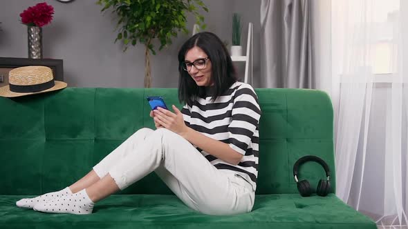 Brunette in Glasses Sitting on Comfortable Sofa at Home and Watching at Her Smartphone