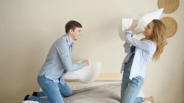 Laughing Couple Fights with Pillows Standing on Knees on Bed