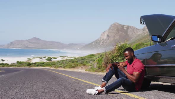 African american man talking on smartphone while standing near his broken down car on road