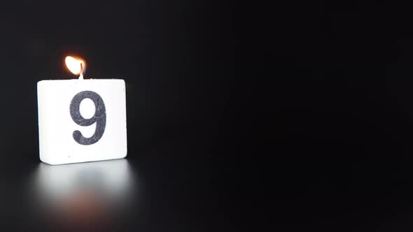 A square candle saying the number 9 being lit and blown out on a dark black background