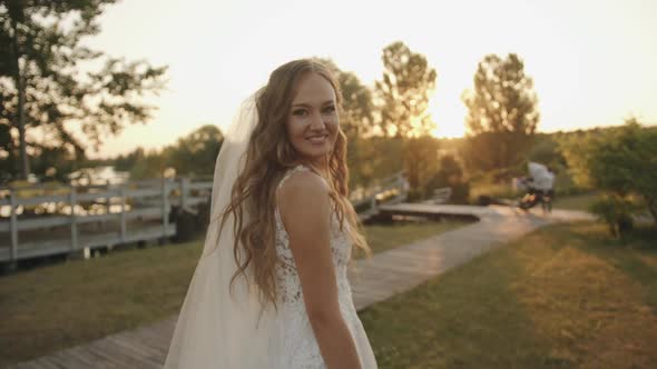 A Spectacular Young Bride Goes to the Wooden Pier on the River Bank at Sunset and Smiles Looking