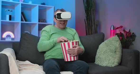 Senior Caucasian Man With a Box of Popcorn in His Hand Watching Video Using Virtual Reality Headset