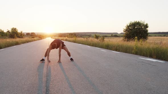 Young Athlete with Sportswear Warming Legs Up on Country Road at Sunset