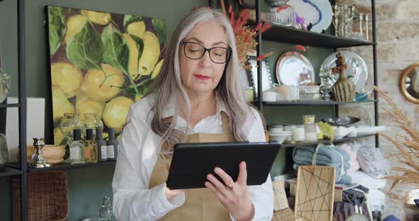 Woman in Glasses which Poses on Camera with Tablet pc while Working on it in Cozy Gift Shop