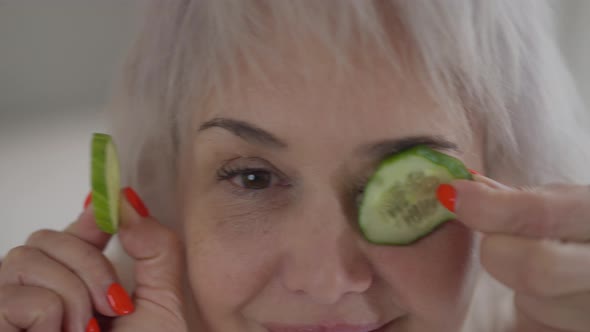 Headshot Cheerful Woman with Cucumber on Eyes Smiling Having Fun Indoors at Home and Eating Slice