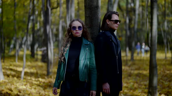 a Man and a Blonde in Leather Jackets and Sunglasses Stand with Their Backs to a Tree in an Autumn