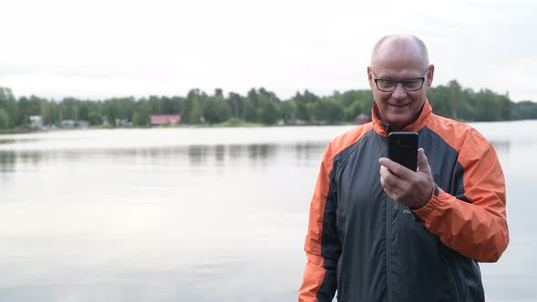 Happy Senior Man Using Phone And Thinking By The Lake