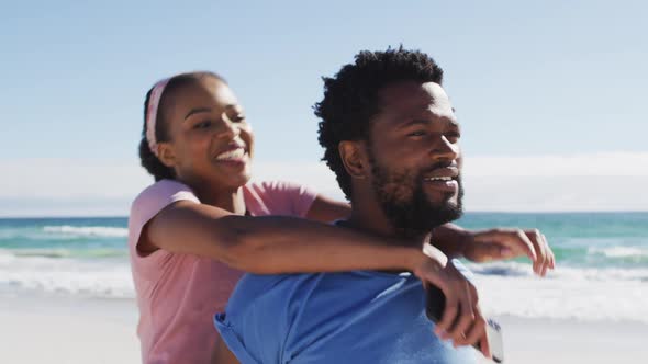 African american couple taking selfie with smartphone, man carrying woman piggyback on the beach