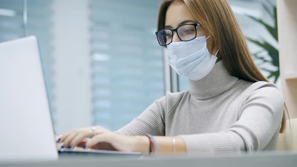 Woman Working at a Laptop in Protective Mask Typing Text in the Office Employee at Work on the