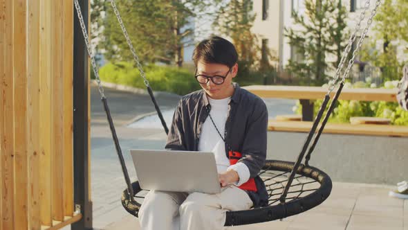 Asian Woman Sitting on Swing Outdoors and Working on Laptop