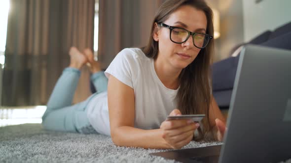 Woman with Glasses Is Lying on the Floor and Makes an Online Purchase Using a Credit Card and Laptop