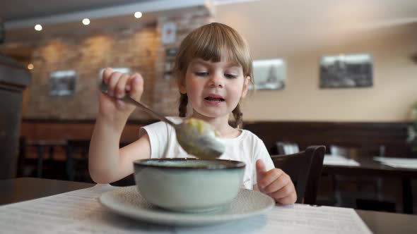 A Small Beautiful Girl with Big Blue Eyes is Eating Soup with a Spoon in a Cafe By Herself