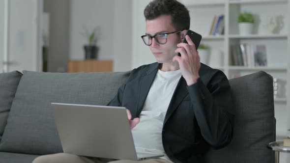 Young Male Designer with Laptop Talking on Smartphone at Home 