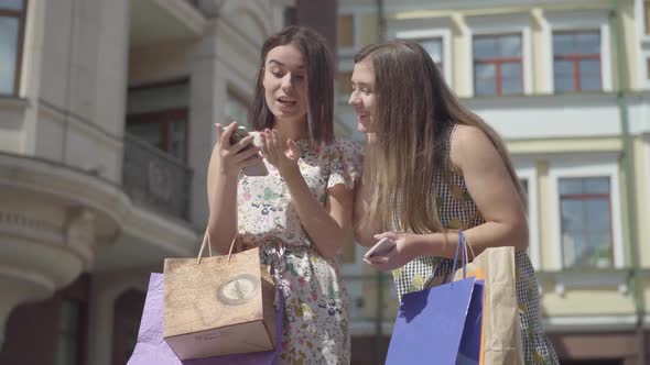 Two Happy Girlfriends After Shopping with Shopping Bags Texting on the Cellphone in Front