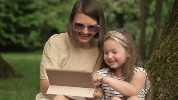 Young Woman with a Little Daughter is Sitting in the Park Under a Large Old Tree and Looking at the