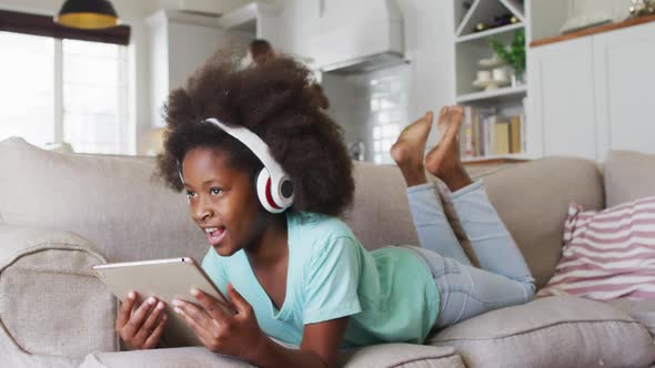 African american girl wearing headphones using tablet lying on couch