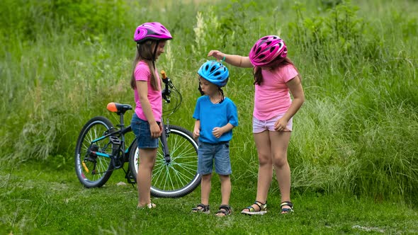 Two girls try to put a protective bicycle helmet on their little brother in a park in the summer