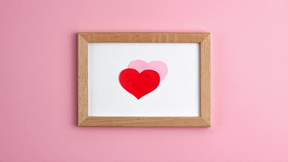 Stop motion animation of pink and red paper hearts in wooden frame on pink background at the centre alt