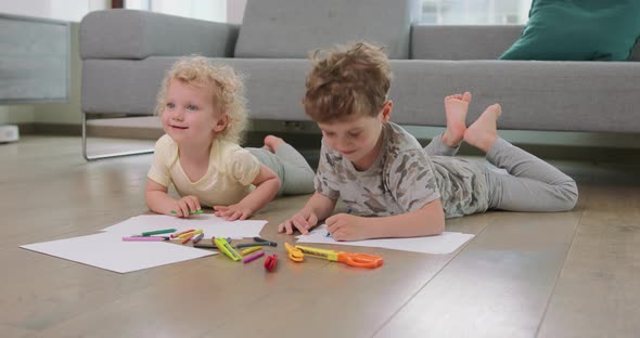 A Little Boy and a Little Girl are Laying on the Floor and Drawing with Pencils on the White Paper