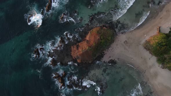 Slowly descending aerial view of a beautiful rocky beach. Small sandy peninsula at sunset. Warm and